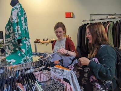 Two people hold clothing on hangars near a circular rack of clothing in a store-like room.